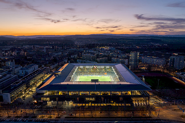 Frontaler Drohnenblick auf das Wankdorfstadion in Bern bei Sonnenuntergang, mit gut sichtbarer Stadionbeleuchtung und der Skyline im Hintergrund.