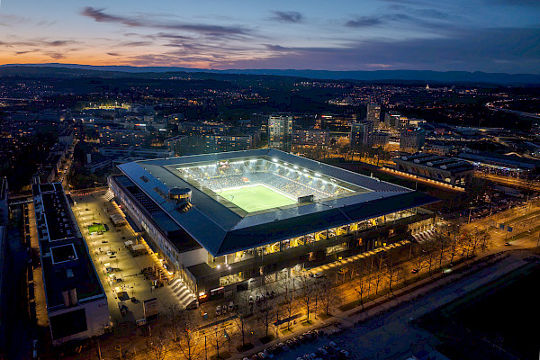 Schräger Drohnenblick auf das hell erleuchtete Wankdorfstadion, eingebettet in die nächtliche Stadt Bern mit Hügeln am Horizont.