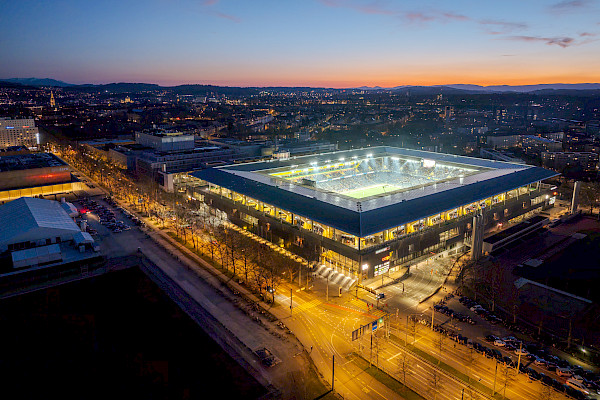 Luftaufnahme des beleuchteten Wankdorfstadions in Bern bei Abenddämmerung, umgeben von Stadtlichtern und Straßenverkehr.